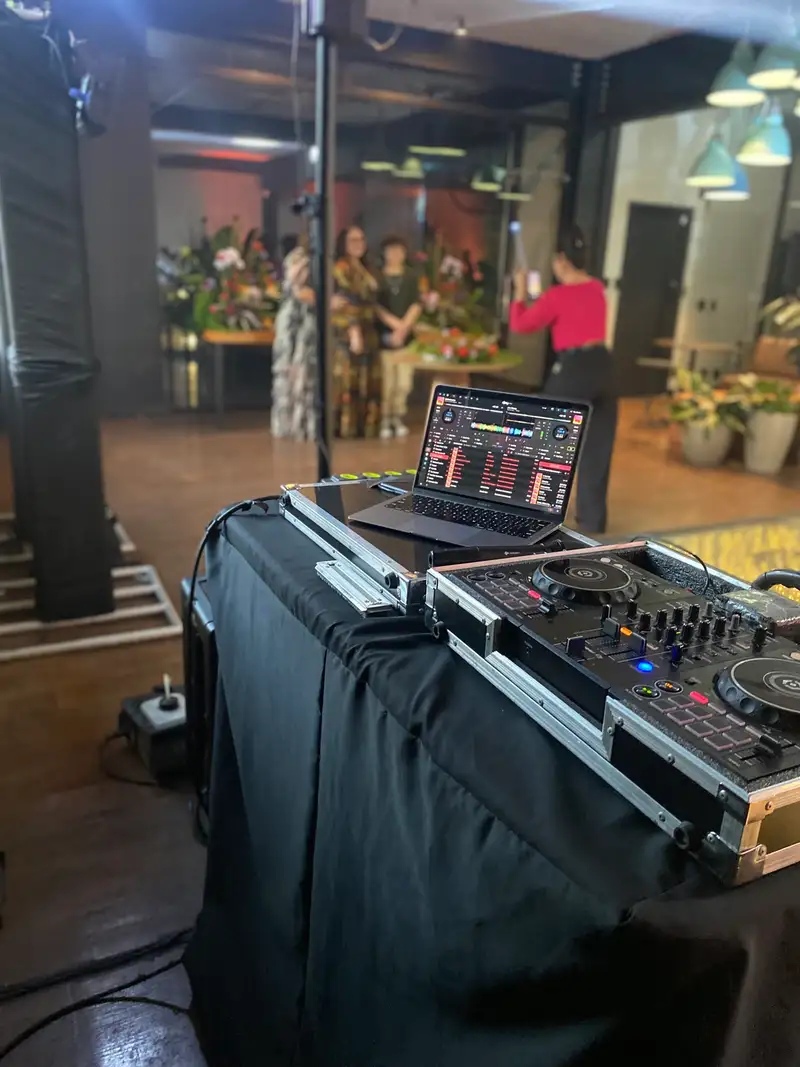 Cabine de DJ com controladora e notebook em primeiro plano, convidados posando para fotos ao fundo