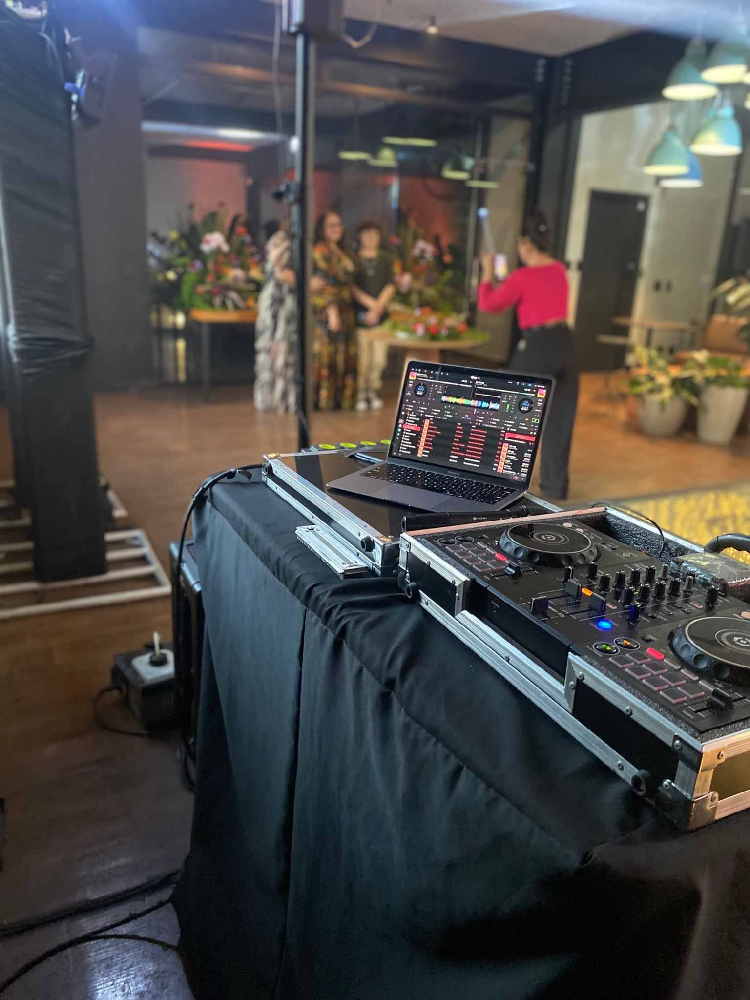 Cabine de DJ com controladora e notebook em primeiro plano, convidados posando para fotos ao fundo