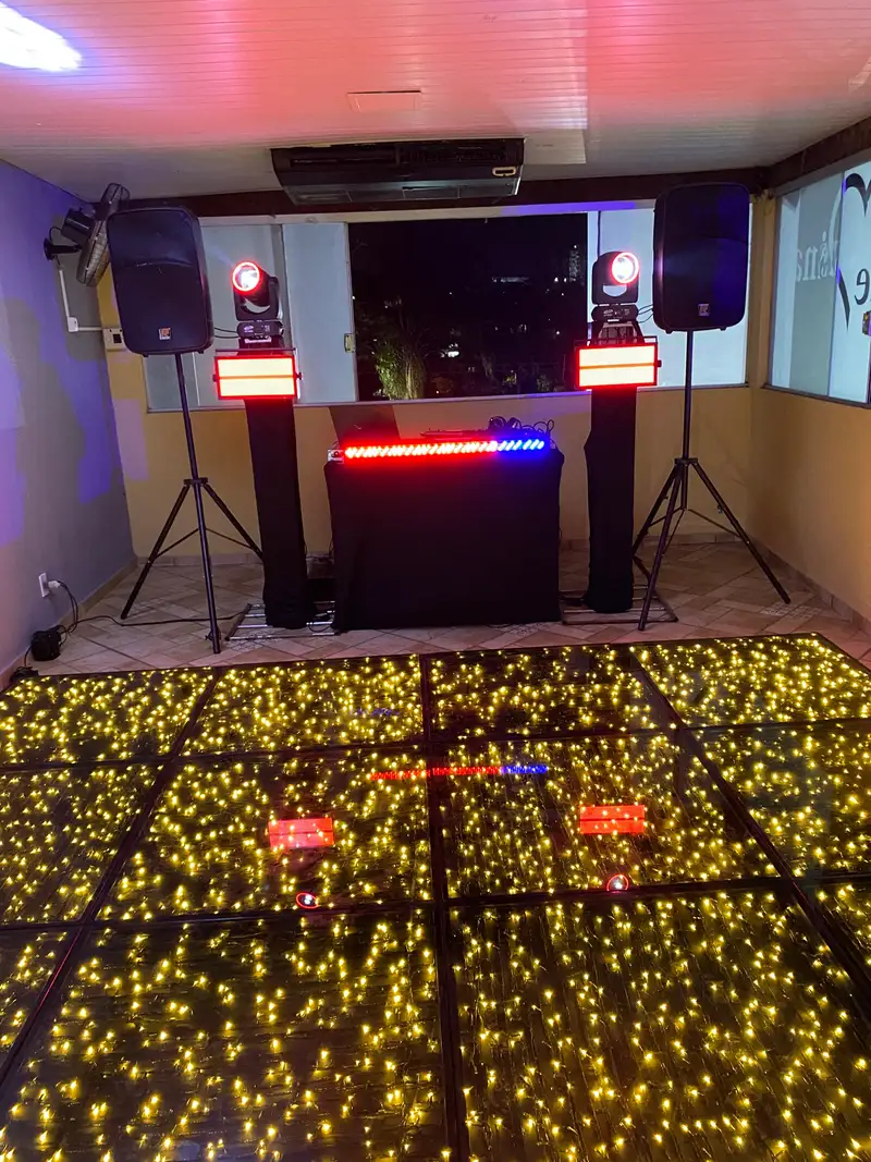 Cabine de DJ com caixas de som, moving heads e strobos diante de uma pista de LED dourada