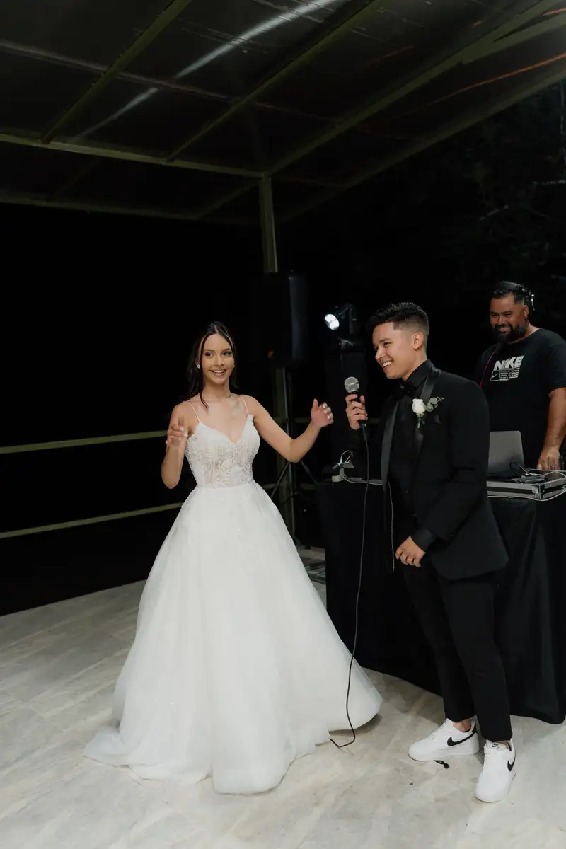 Noiva sorrindo ao lado de um convidado ao microfone, com caixa de som e moving head ao fundo