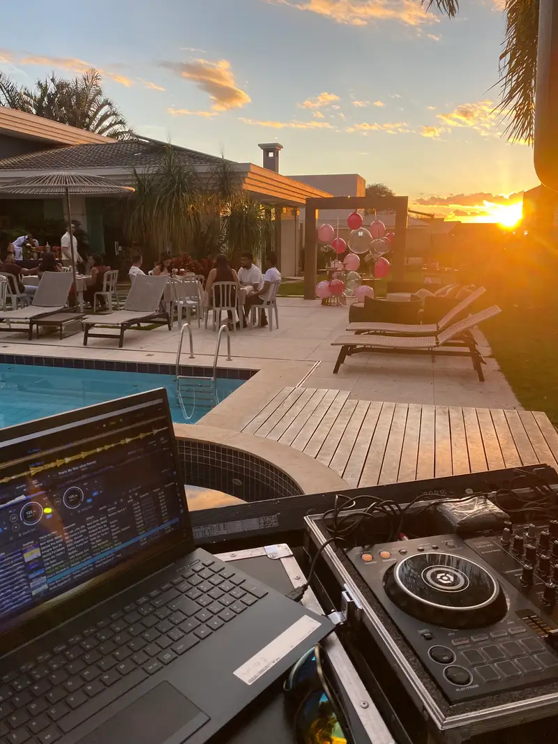 Cabine de DJ à beira da piscina ao pôr do sol, com convidados à mesa no jardim de uma casa