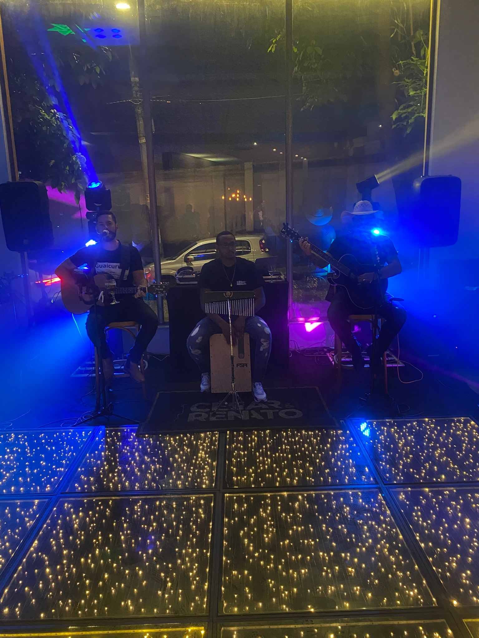 Trio acústico tocando atrás de uma pista de LED, com luz azul e violeta
