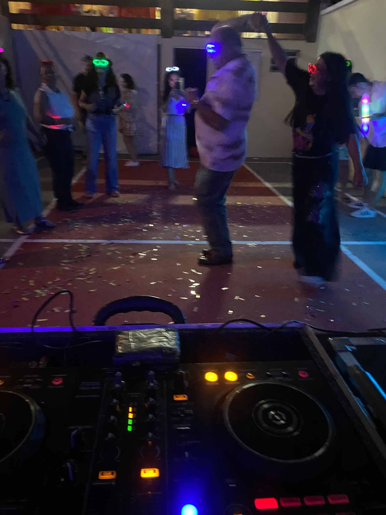 Convidados dançando com óculos de LED na pista, vistos da cabine do DJ