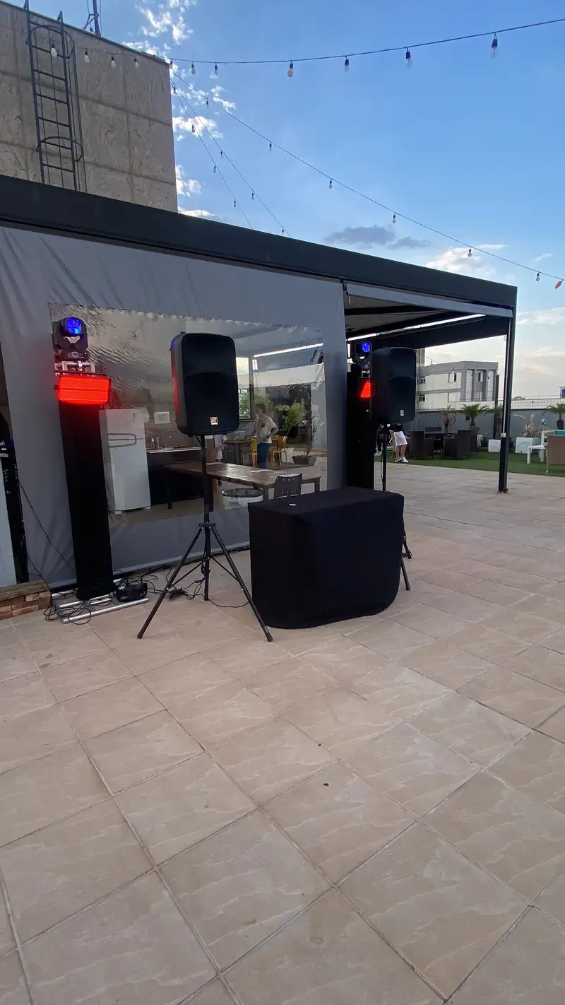 Caixas de som em tripé, moving heads e cabine de DJ montados em uma cobertura, à tarde