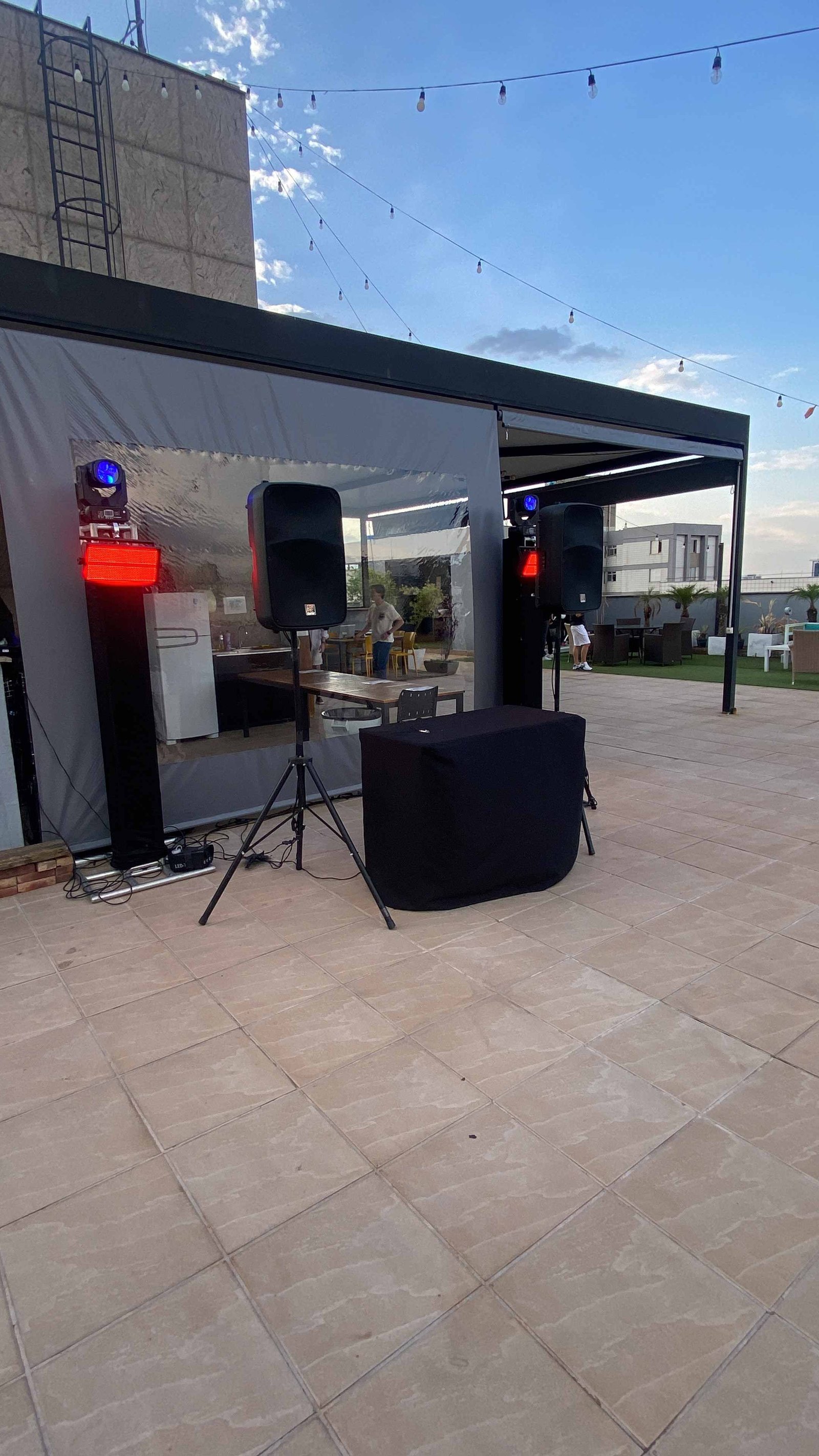 Caixas de som, moving heads e cabine de DJ montadas no terraço de uma cobertura, sob varal de luzes